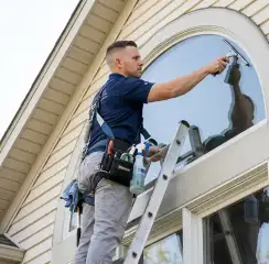 Professional window cleaner on a ladder using a squeegee to clean an exterior arched window, Tulsa, OK.